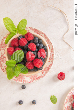 A high angle shot displays a rustic bowl brimming with fresh raspberries and blueberries decorated with mint sprigs on a floral vintage plate over a bright textured surface with scattered fruit 140860021