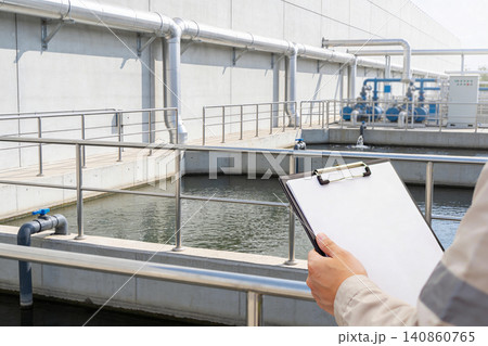 Worker inspects water treatment plant with clipboard, monitoring industrial processes Worker inspects water treatment plant with clipboard, monitoring industrial processes 140860765