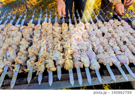Two hands are holding skewers with meat grilling on an open flame outside in daylight, giving view of many skewers. Two hands are holding skewers with meat grilling on an open flame outside in daylight, giving view of many skewers. 140862789