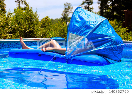 Woman relaxes on floating lounge in pool with canopy, surrounded by green trees, enjoying sunny day. Woman relaxes on floating lounge in pool with canopy, surrounded by green trees, enjoying sunny day. 140862790