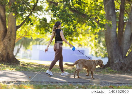 Young woman walking a golden retriever dog on a sunny day in a park with green trees and a paved pathway surrounded by autumn leaves 140862909