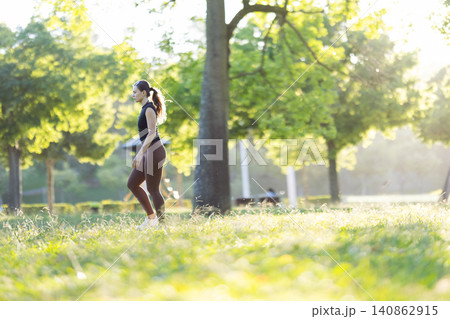 Female jogger in black athletic wear walks through a sunlit park with green trees and grass, enjoying a healthy lifestyle in a natural outdoor setting 140862915