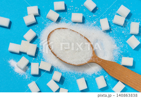 A wooden spoon with a mountain of white granulated sugar on a blue background with scattered crystals and cubes of refined sugar. A wooden spoon with a mountain of white granulated sugar on a blue background with scattered crystals and cubes of refined sugar. 140863838