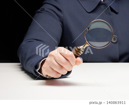 A woman's hand in a blue jacket holds an antique magnifying glass over a white table A woman's hand in a blue jacket holds an antique magnifying glass over a white table 140863911