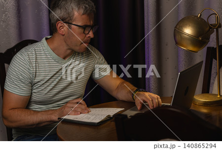 Man working at home desk with laptop and notebook during evening hours Man working at home desk with laptop and notebook during evening hours 140865294