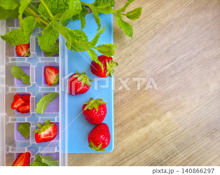 Preparing ice cubes with fresh strawberries and mint leaves in a plastic tray on a wooden table. High angle view. Summer refreshing drinks ingredients. Fresh strawberry and green mint in ice cube tray Preparing ice cubes with fresh strawberries and mint leaves in a plastic tray on a wooden table. High angle view. Summer refreshing drinks ingredients. Fresh strawberry and green mint in ice cube tray 140866297