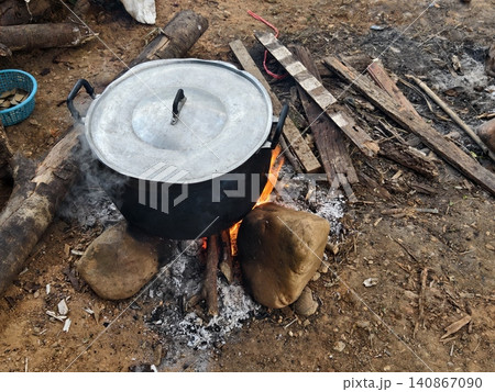 A high-angle shot of a large aluminium cooking pot boiling over an open wood fire supported by stones. Surrounded by scattered firewood, representing outdoor camping and rural cooking methods. A high-angle shot of a large aluminium cooking pot boiling over an open wood fire supported by stones. Surrounded by scattered firewood, representing outdoor camping and rural cooking methods. 140867090