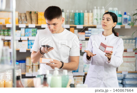 Handsome young man choosing box with pills in pharmacy, scan QR code on label and professional pharmacist woman helping and offering supplements at drugstore Handsome young man choosing box with pills in pharmacy, scan QR code on label and professional pharmacist woman helping and offering supplements at drugstore 140867664