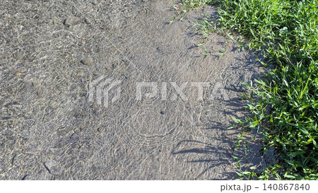 A horizontal high angle perspective captures clear freshwater flowing gently over a shallow sandy riverbed bordered by lush green grass. Bright afternoon sunlight creates intricate shimmering patterns 140867840