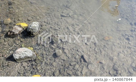 A horizontal high angle overhead perspective captures clear freshwater flowing over a shallow rocky riverbed transitioning into cloudy, murky water sediment. Bright afternoon sunlight creates fine 140867844