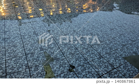 High angle full frame shot captures ripples on clear water flowing over a gray stone mosaic tiled floor, displaying shimmering golden light reflections from decorative evening illumination. Modern High angle full frame shot captures ripples on clear water flowing over a gray stone mosaic tiled floor, displaying shimmering golden light reflections from decorative evening illumination. Modern 140867861