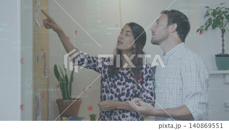 Coworkers pointing and holding notepad at corkboard with sketches in office nook, in casual shirts Coworkers pointing and holding notepad at corkboard with sketches in office nook, in casual shirts 140869551