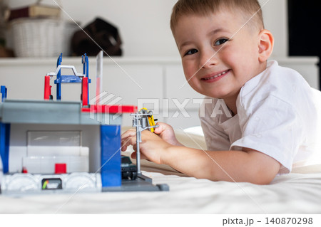 Cute young boy lying on a white blanket and focusing on playing with a toy garage, car repair set, and colorful scale model vehicles. 140870298