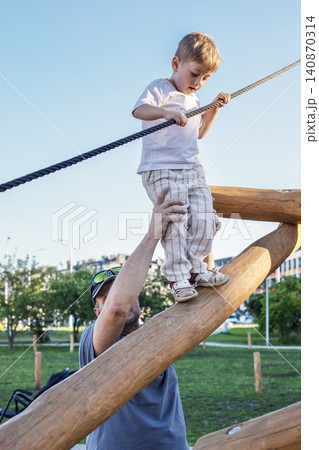 A caring father hands helps his little toddler boy walking safely on a wooden logs balance structure at outdoor city playground. A caring father hands helps his little toddler boy walking safely on a wooden logs balance structure at outdoor city playground. 140870314
