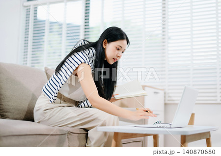 Young Asian woman writing notes in a notebook while using a laptop at home. Remote work, online learning, productivity and modern lifestyle concept. Young Asian woman writing notes in a notebook while using a laptop at home. Remote work, online learning, productivity and modern lifestyle concept. 140870680