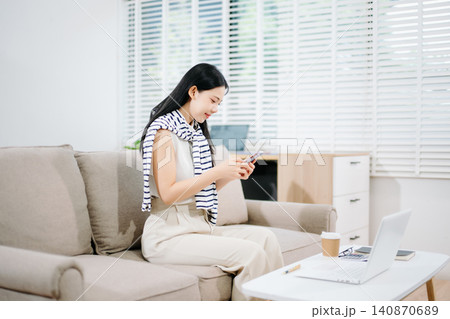 Happy Asian woman analyzing financial data documents and working on her laptop while sitting comfortably on a living room sofa. Happy Asian woman analyzing financial data documents and working on her laptop while sitting comfortably on a living room sofa. 140870689