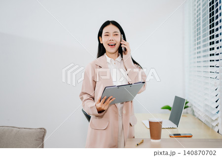 Happy Asian Businesswoman Reviewing Documents and Talking on Smartphone by Office Window in Modern Home Workspace 140870702