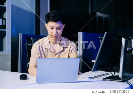 Happy Asian young man smiling while typing on his laptop at a modern corporate office desk at night. Happy Asian young man smiling while typing on his laptop at a modern corporate office desk at night. 140871092