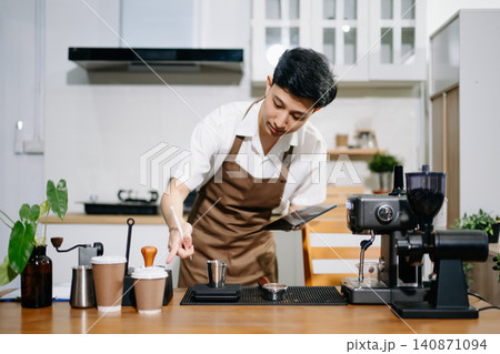 Male Asian barista using an espresso machine and taking notes on a clipboard in coffee shop. 140871094