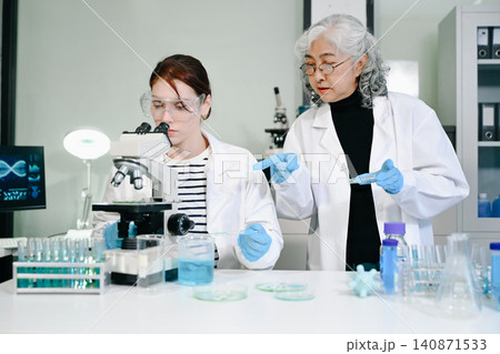 Two diverse female scientists analyze biological samples in a modern lab, utilizing a microscope and petri dishes 140871533