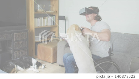 Smiling man in wheelchair with VR headset and jeans holding controller with Labrador in living room Smiling man in wheelchair with VR headset and jeans holding controller with Labrador in living room 140875322