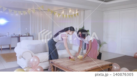 Leaning two women arranging snacks on living room table, placing glass bowl, wearing casual clothes Leaning two women arranging snacks on living room table, placing glass bowl, wearing casual clothes 140876305