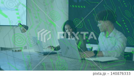 Discussing woman wearing blazer and man wearing shirt using laptops at office table, green overlay Discussing woman wearing blazer and man wearing shirt using laptops at office table, green overlay 140876333