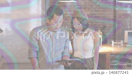 Discussing colleagues leaning over tablet in loft, wearing sleeves rolled, beanie, star overlay Discussing colleagues leaning over tablet in loft, wearing sleeves rolled, beanie, star overlay 140877241
