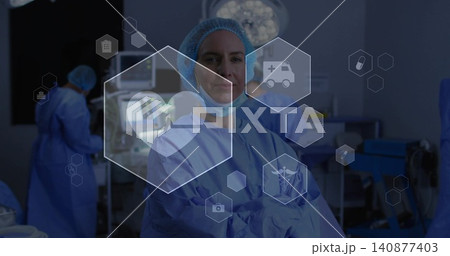 Standing surgeon in OR wearing blue gown, bouffant cap, mask lowered, crossing arms, showing hex UI 140877403