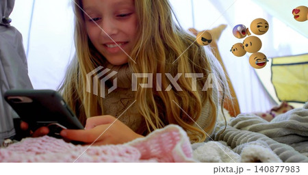 Smiling girl lying on stomach in tent, wearing tan sweater and holding smartphone with emoji Smiling girl lying on stomach in tent, wearing tan sweater and holding smartphone with emoji 140877983