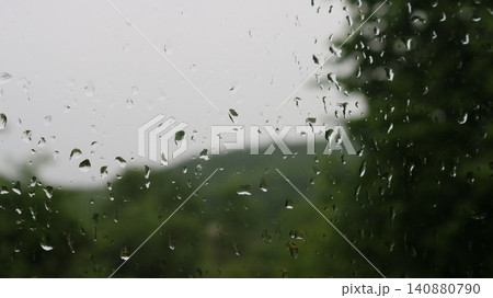 Large raindrops on a windowpane. Through the wet surface, a very blurred silhouette of a green landscape with hills and trees under an overcast sky is visible, creating a melancholy atmosphere. 140880790
