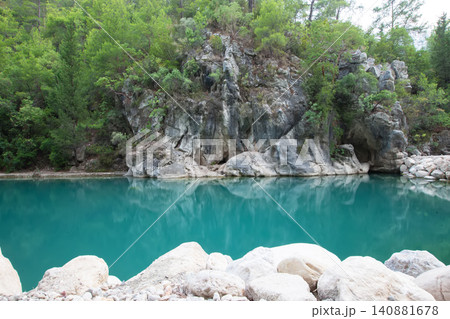 Clear turquoise lake surrounded by lush greenery and rocky cliffs during a sunny day in a tranquil natural setting Clear turquoise lake surrounded by lush greenery and rocky cliffs during a sunny day in a tranquil natural setting 140881678
