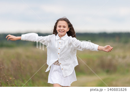 Smiling girl in white outfit joyfully running through a green field on a cloudy day Smiling girl in white outfit joyfully running through a green field on a cloudy day 140881826