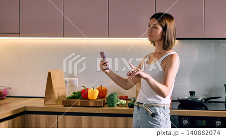 Woman using smartphone to manage grocery order in kitchen. 140882074