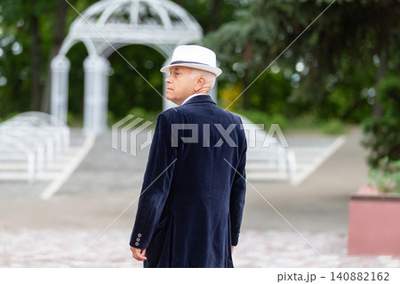 Elderly man in stylish coat and hat walks through park with gazebo in the background during daytime Elderly man in stylish coat and hat walks through park with gazebo in the background during daytime 140882162