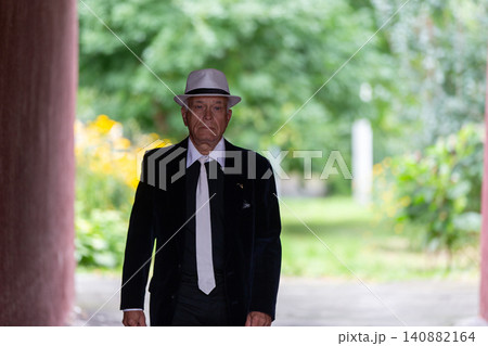 Elderly gentleman walks through garden pathway dressed in formal attire with a hat on a sunny day Elderly gentleman walks through garden pathway dressed in formal attire with a hat on a sunny day 140882164