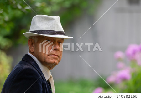 Older man wearing a stylish hat looks thoughtfully at the camera in a vibrant garden setting during a sunny day 140882169
