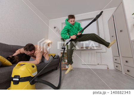 The child jumps joyfully during the vacuum cleaner as a friend plays with a pets in a cozy living room during a daytime event The child jumps joyfully during the vacuum cleaner as a friend plays with a pets in a cozy living room during a daytime event 140882343