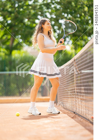 Tennis player practices on court during sunny day with green trees in background Tennis player practices on court during sunny day with green trees in background 140884551