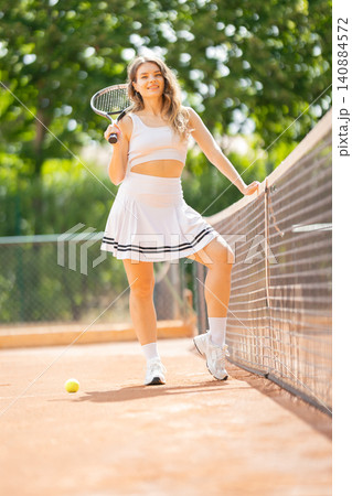 Woman stands on tennis court holding racket with one leg on the net and a ball on the ground during warm sunny day Woman stands on tennis court holding racket with one leg on the net and a ball on the ground during warm sunny day 140884572