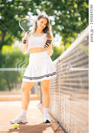 Young woman stands on a tennis court holding a racket and checking her phone during a sunny afternoon practice session 140884590