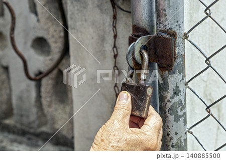 Close Up of Elderly Wrinkled Hand Holding and Unlocking an Old Rusty Padlock Secured to a Weathered Metal Gate Post With Chain Link Fence and Aged Concrete Wall Background in Rustic Outdoor Setting Close Up of Elderly Wrinkled Hand Holding and Unlocking an Old Rusty Padlock Secured to a Weathered Metal Gate Post With Chain Link Fence and Aged Concrete Wall Background in Rustic Outdoor Setting 140885500