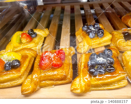 Assorted puff pastry danishes topped with fresh strawberries, blueberries, and kiwi slices on wooden bakery display slats. Delicious glazed fruit tarts and sweet desserts for sale in a cafe. 140888157