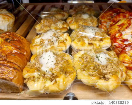 Assorted savory pastries displayed on wooden shelves in a bakery. Featuring buns topped with pork floss, mayonnaise, and cheese, alongside pizza bread and herbal loaves. Assorted savory pastries displayed on wooden shelves in a bakery. Featuring buns topped with pork floss, mayonnaise, and cheese, alongside pizza bread and herbal loaves. 140888259