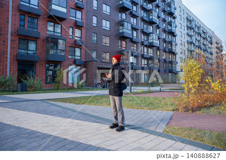 Man standing in modern residential courtyard with smartphone. Hipster guy in red hat using mobile phone near apartment building. Urban lifestyle and digital city communication concept. Man standing in modern residential courtyard with smartphone. Hipster guy in red hat using mobile phone near apartment building. Urban lifestyle and digital city communication concept. 140888627