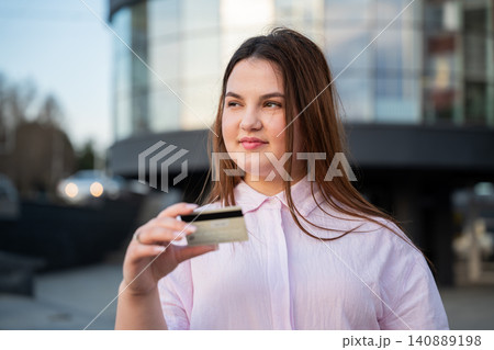 Young woman holding a bank card outside a modern office building during the workday. Online payment and secure digital transactions for everyday cashless shopping Young woman holding a bank card outside a modern office building during the workday. Online payment and secure digital transactions for everyday cashless shopping 140889198