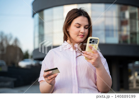 Young woman using a smartphone and holding a bank card outside a modern office building during the workday. Mobile banking and digital banking for secure everyday financial management Young woman using a smartphone and holding a bank card outside a modern office building during the workday. Mobile banking and digital banking for secure everyday financial management 140889199