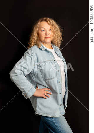 Woman 57 years old with curly hair wearing denim shirt on black background. Portrait of attractive lady looking at camera. Fashionable casual style for mature person lifestyle photography. 140889959