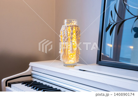 Macrame lamp with fairy light in glass vase. Handmade home decor item sitting on white piano. Interior design accessory for cozy atmosphere and soft warm glow in living room living space. Macrame lamp with fairy light in glass vase. Handmade home decor item sitting on white piano. Interior design accessory for cozy atmosphere and soft warm glow in living room living space. 140890174