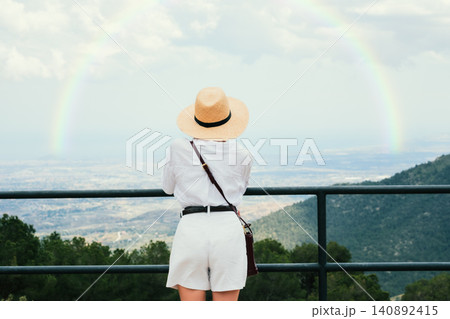 Woman in straw hat admiring rainbow from mountain viewpoint on a cloudy day Woman in straw hat admiring rainbow from mountain viewpoint on a cloudy day 140892415